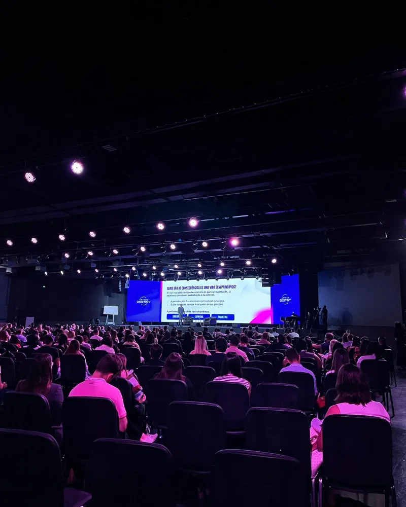 Conferência na Casa de Destino com plateia cheia, telões de LED no palco e luzes no teto