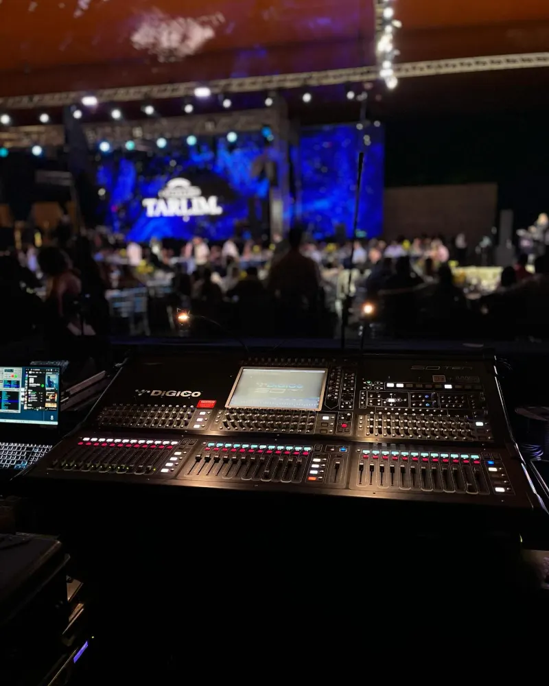 Mesa de som na técnica, com o palco iluminado e o público ao fundo
