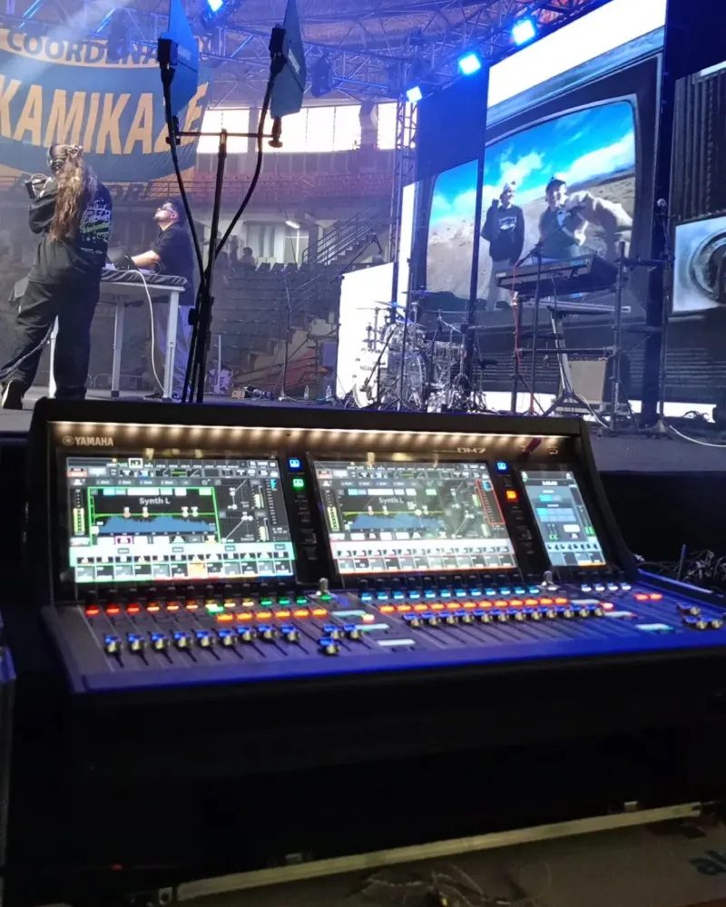 Duas mesas de som digitais na técnica de uma arena, com o palco e os telões ao fundo